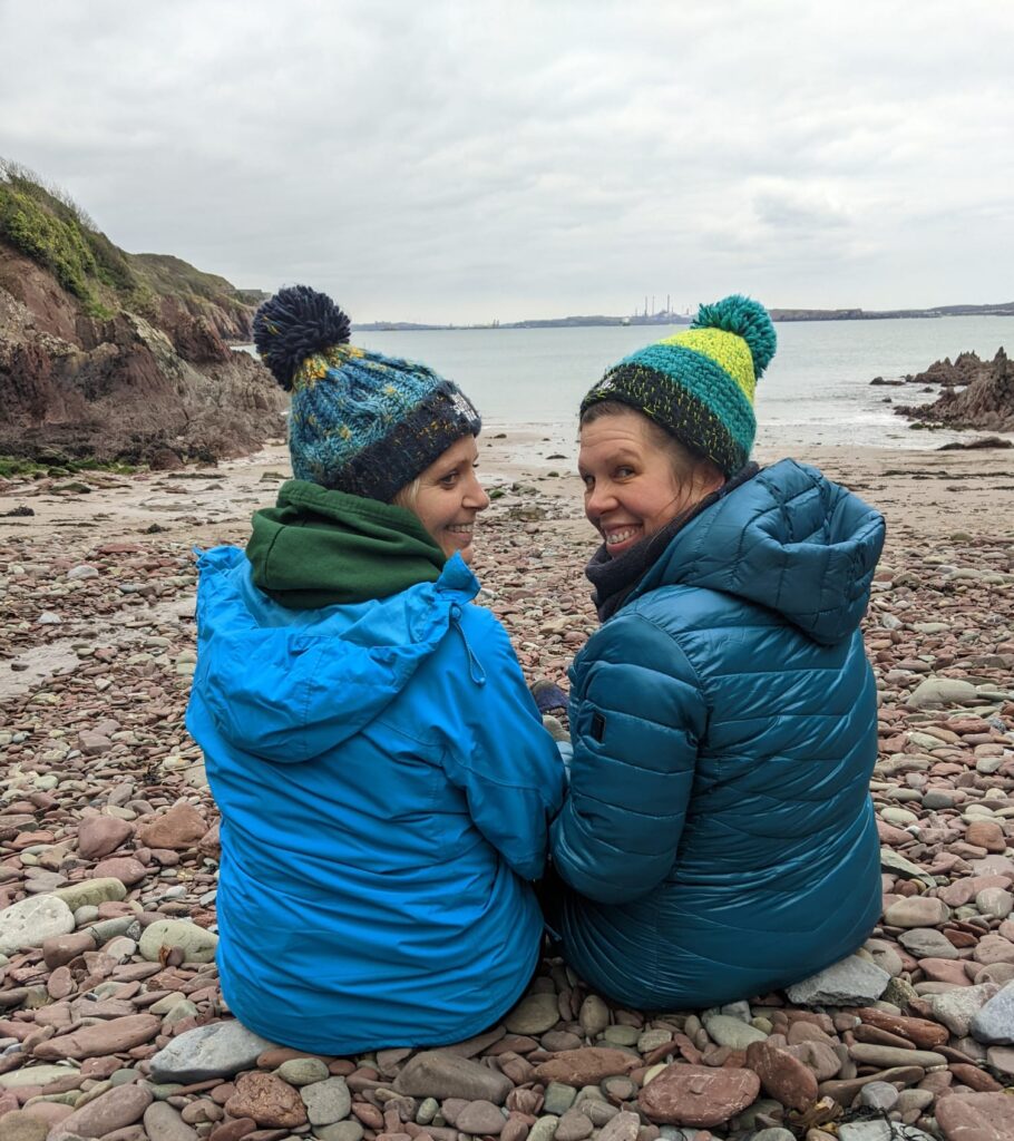 2 smiling friends wearing woolly hats sat on a pebbly beach enjoying a Wild swim & yoga retreat in Pembrokeshire